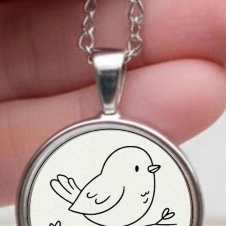 Silver necklace with a circular pendant displaying a black and white drawing of a bird on a branch. The pendant is held in a hand against a white backdrop.