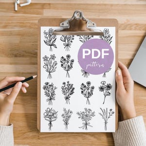 Può includere: Un fermacarte contiene un foglio bianco con disegni floreali neri. Un cerchio viola con il testo "PDF pattern" è sul foglio. Una mano tiene una matita e l'altra tiene il fermacarte. Lo sfondo è una superficie di legno.
