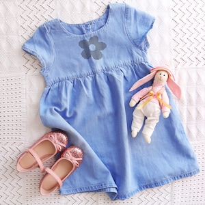 Può includere: Un vestito in denim azzurro con un motivo floreale nero, abbinato a scarpe rosa glitterate e un coniglietto di peluche con cappello e giacca. L'abito ha maniche corte e vita arricciata.