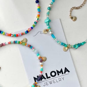 Può includere: Collane di perline colorate con ciondoli a forma di conchiglia dorata. Le collane presentano una varietà di perline nei colori dell'arcobaleno, turchese e bianco. Le collane sono esposte su una superficie bianca con un biglietto bianco che dice "NALOMA JEWELRY".