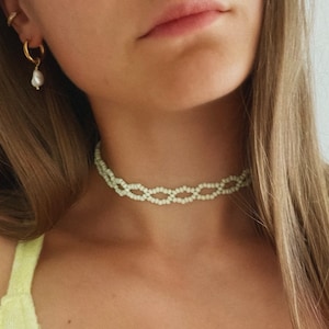 Può includere: Un girocollo di perline verde chiaro con un design intrecciato. La collana è indossata da una persona con i capelli lunghi e castani.
