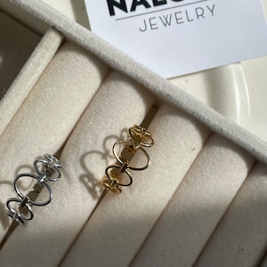 Könnte beinhalten: Zwei goldene und silberne Ringe mit einem einzigartigen, ineinandergreifenden Design. Die Ringe werden in einer weißen Samt-Ringbox präsentiert. Die Box trägt ein Etikett mit der Aufschrift "NALOMA JEWELRY".