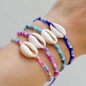 Può includere: Cinque braccialetti colorati con perline e ciondoli a forma di conchiglia cauri. I braccialetti sono in tonalità di rosa, viola, blu e turchese.