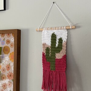 May include: A pink, green, and white crocheted wall hanging with a cactus design and fringe. The hanging is attached to a wooden dowel rod and white string.