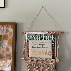 May include: A pink crocheted wall hanging with two wooden dowels, holding a magazine and a book titled "Atomic: Tiny Changes, Remarkable Results".
