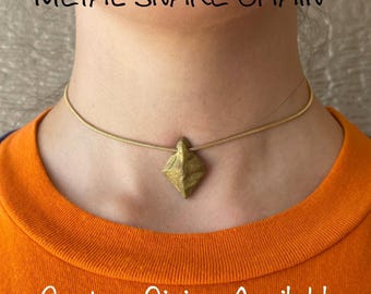 May include: A gold-coloured metal snake chain necklace with a geometric pendant. The necklace is worn on a person with an orange top. Text at the top reads "METAL SNAKE CHAIN" and at the bottom "Custom Sizing Available".