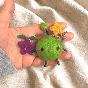 May include: A selection of handmade keyrings with quirky designs. The keyrings include a green felt creature with black legs and arms, a purple star with green wings, and a yellow star. The keyrings are attached to a silver chain.