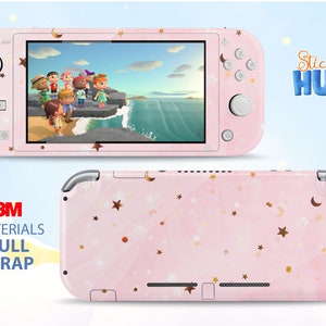 Op de afbeelding: Een roze Nintendo Switch Lite console skin met een patroon van gouden sterren en manen. Het scherm toont een videogame scène. De woorden "Sticky HUG" en "3M Materials Full Wrap" zijn ook zichtbaar.