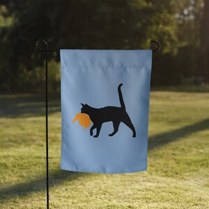 May include: A light blue garden flag featuring a black cat silhouette carrying an orange object. The flag is hanging on a black metal pole in a grassy outdoor setting.