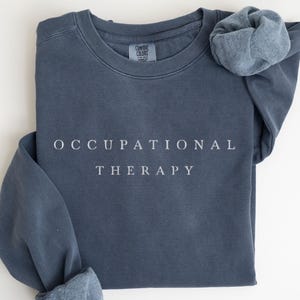 May include: Denim blue sweatshirt with the words "OCCUPATIONAL THERAPY" printed in white. The sweatshirt has a crew neck and long sleeves with cuffed wrists. The word "DENIM" is printed in black at the bottom.