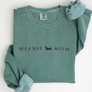 Puede incluir: Sudadera verde claro con el texto "WEENIE MOM" y una ilustraci&oacute;n de perro salchicha. La sudadera tiene cuello redondo y mangas largas. La parte inferior de la imagen tiene el texto "LIGHT GREEN".