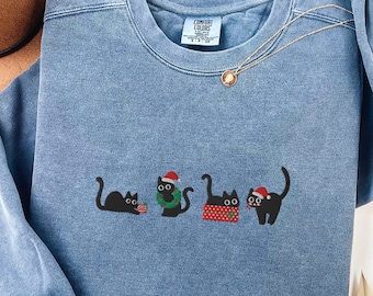 May include: Blue jean-colored sweatshirt with a crew neck and long sleeves. The sweatshirt features an embroidered design of four black cats, each wearing a festive accessory. The text "BLUE JEAN" is at the bottom.
