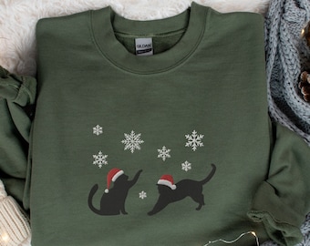 May include: A green Christmas sweatshirt with two black cats wearing Santa hats and playing in the snow. The sweatshirt has a snowflake design.