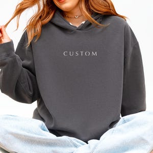 May include: A dark gray hoodie with the word "CUSTOM" embroidered on the front. The hoodie has a drawstring hood and long sleeves. The word "PEPPER" is printed at the bottom right corner of the image.