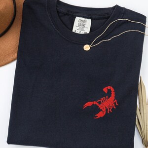May include: A black crew neck t-shirt featuring a small red embroidered scorpion on the lower right side. The inner neck label reads Comfort Colors and indicates size S-P-CH. The shirt is displayed with a brown hat and a gold necklace.