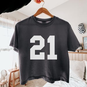 May include: Dark gray t-shirt with the number 21 in white block letters. The shirt is hanging on a wooden hanger. The shirt is made of a soft material.