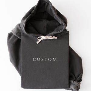 May include: A black hoodie with a drawstring hood and the word "CUSTOM" printed in white. The word "BLACK" is printed in white at the bottom right corner. The hoodie is folded on a white surface.
