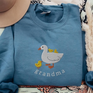 May include: A blue sweatshirt with a white embroidered goose and three yellow ducklings. The text "grandma" is embroidered below the goose.