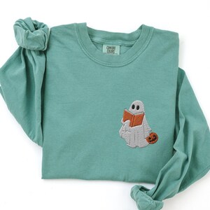 K&ouml;nnte beinhalten: Tealfarbenes Langarmshirt mit einem gestickten Geist, der ein Buch und einen K&uuml;rbis h&auml;lt. Das Shirt hat den Text "Comfort Colors" auf dem Etikett.