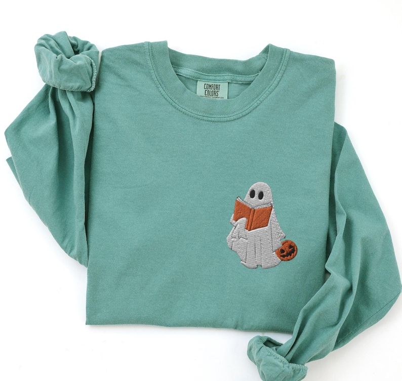 K&ouml;nnte beinhalten: Tealfarbenes Langarmshirt mit einem gestickten Geist, der ein Buch und einen K&uuml;rbis h&auml;lt. Das Shirt hat den Text "Comfort Colors" auf dem Etikett.