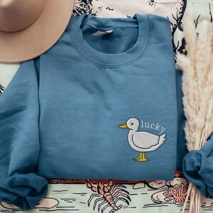 Puede incluir: Una sudadera azul con un pato bordado en blanco y la palabra "lucky" en la parte delantera.