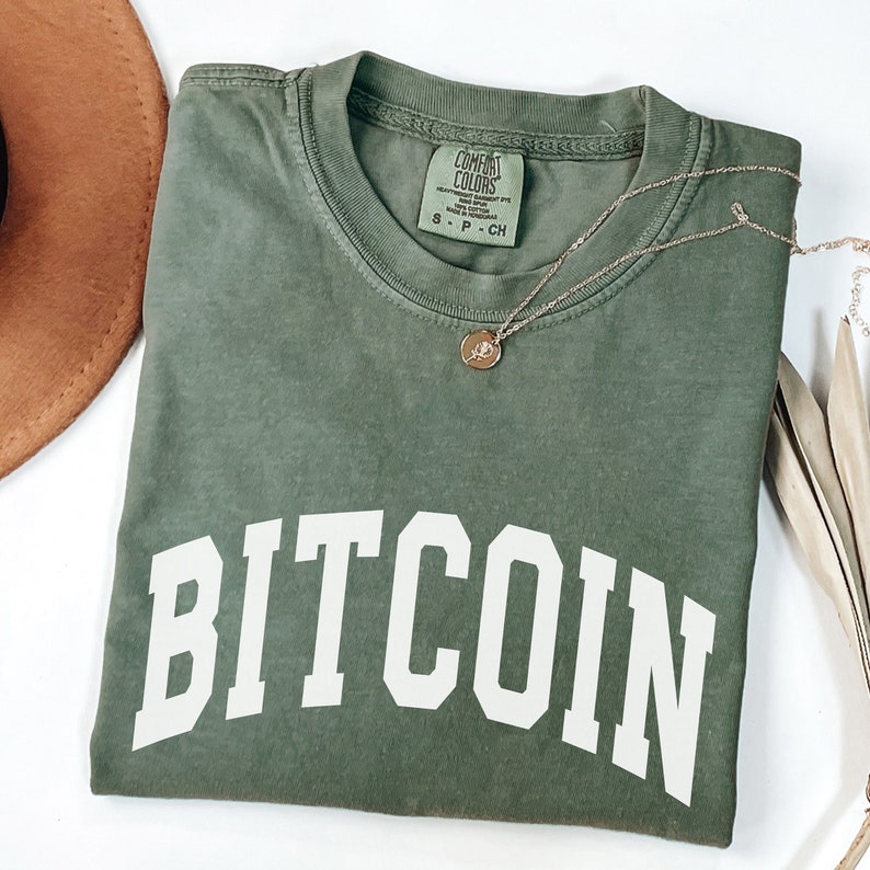 May include: A green t-shirt with the word "BITCOIN" printed in white on the front. The shirt is made of a soft, comfortable material and has a relaxed fit.