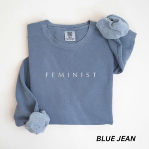 Pu&ograve; includere: Felpa blu jeans con la scritta "FEMINIST" stampata in bianco. La felpa ha un girocollo e maniche lunghe, con uno scrunchie abbinato sopra. La parte inferiore dell'immagine recita "BLUE JEAN".