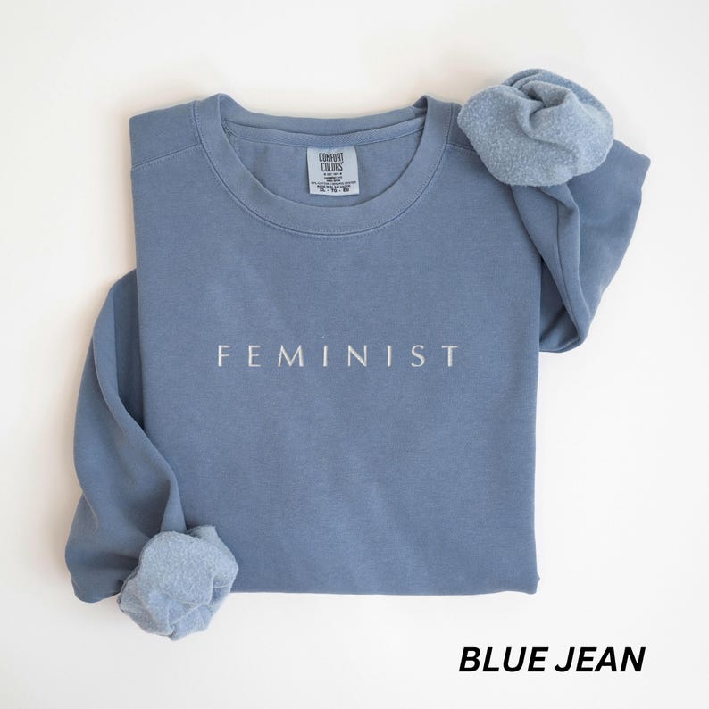 Pu&ograve; includere: Felpa blu jeans con la scritta "FEMINIST" stampata in bianco. La felpa ha un girocollo e maniche lunghe, con uno scrunchie abbinato sopra. La parte inferiore dell'immagine recita "BLUE JEAN".