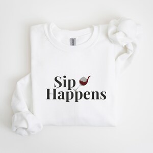 May include: White sweatshirt with the text "Sip Happens" and a graphic of a wine glass with red wine spilling out.
