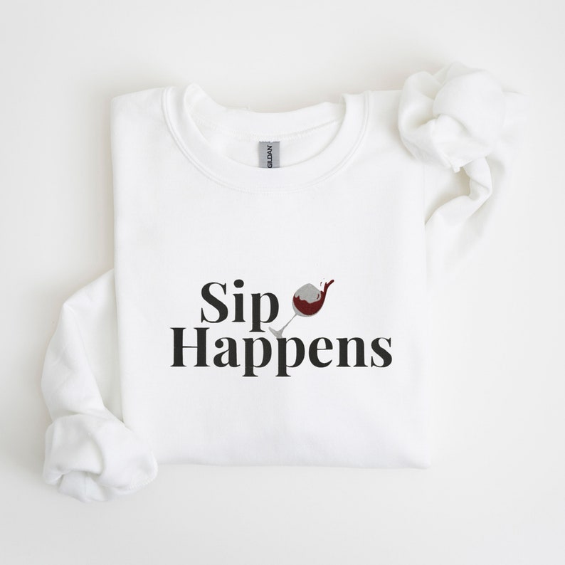 May include: White sweatshirt with the text "Sip Happens" and a graphic of a wine glass with red wine spilling out.