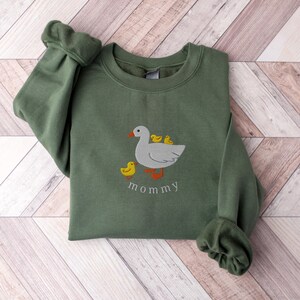 May include: A green sweatshirt with an embroidered image of a white duck with three yellow ducklings. The text "mommy" is embroidered below the duck family.