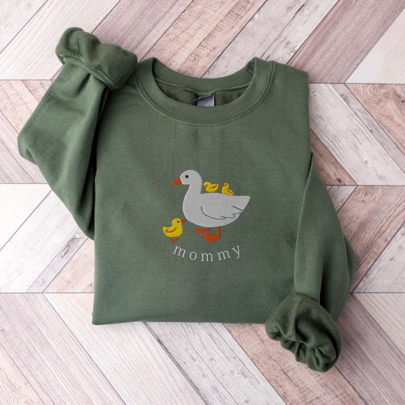 May include: A green sweatshirt with an embroidered image of a white duck with three yellow ducklings. The text "mommy" is embroidered below the duck family.