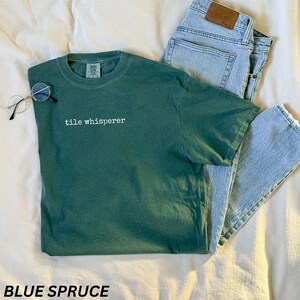K&ouml;nnte beinhalten: Ein blau-fichtefarbenes T-Shirt mit dem wei&szlig;en Text "tile whisperer", kombiniert mit hellblauen Jeans und runden Brillen. Das T-Shirt ist einfarbig und hat einen Rundhalsausschnitt.