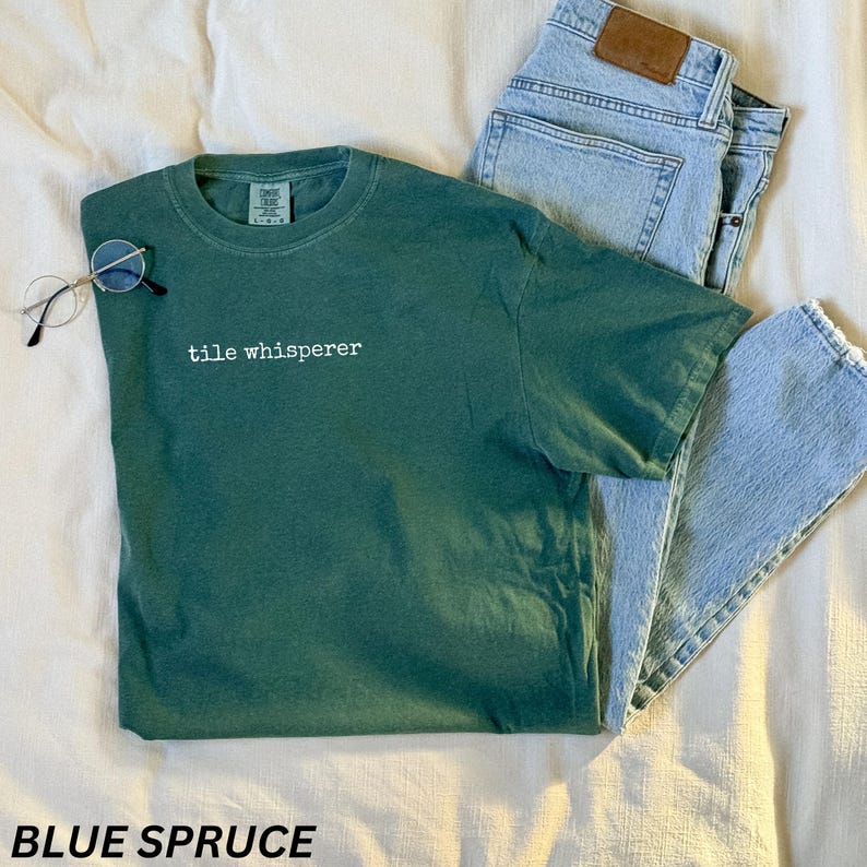 K&ouml;nnte beinhalten: Ein blau-fichtefarbenes T-Shirt mit dem wei&szlig;en Text "tile whisperer", kombiniert mit hellblauen Jeans und runden Brillen. Das T-Shirt ist einfarbig und hat einen Rundhalsausschnitt.