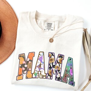 Puede incluir: Una camiseta de color crema con la palabra "MAMA" impresa en letras grandes. Cada letra est&aacute; llena de un patr&oacute;n diferente con tem&aacute;tica de Halloween, que incluye calabazas, fantasmas, ara&ntilde;as y girasoles.