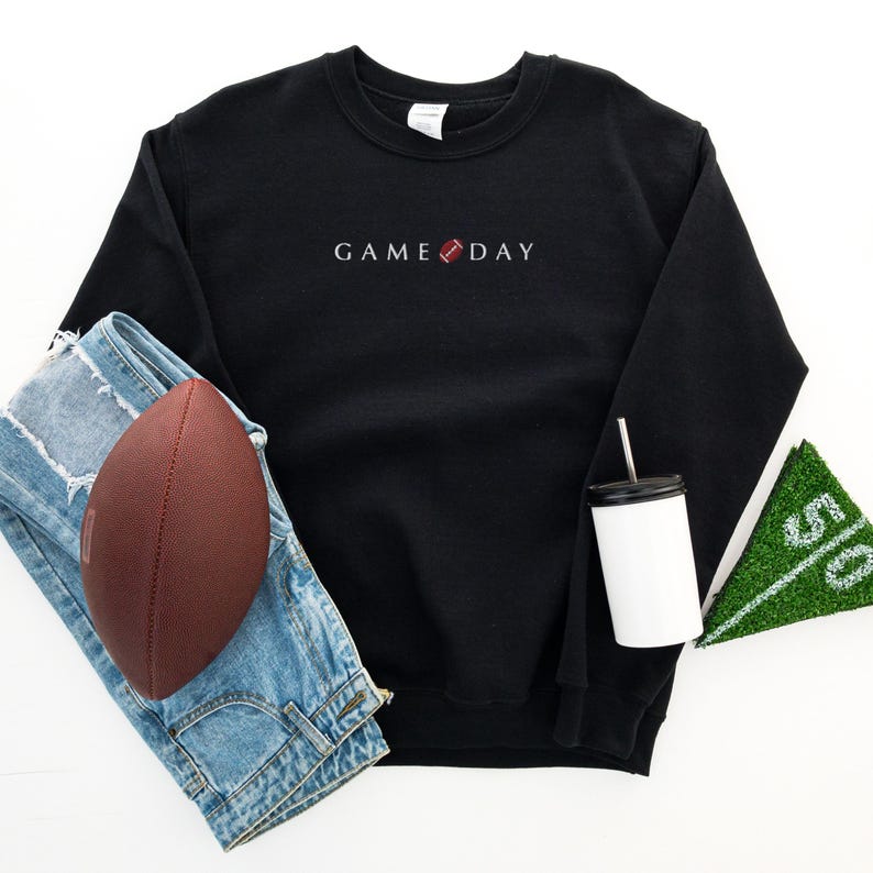 Może przedstawiać: Czarna bluza z napisem "GAME DAY" i grafiką piłki nożnej. Obraz zawiera brązową piłkę nożną, niebieskie dżinsy, biały kubek z czarną pokrywką i słomką oraz zielony znacznik pola.