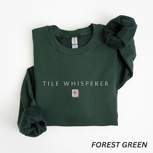 May include: Forest green sweatshirt with the text "TILE WHISPERER" in white. Below the text is a small square graphic with a red and gold design. The sweatshirt is folded with the sleeves visible.