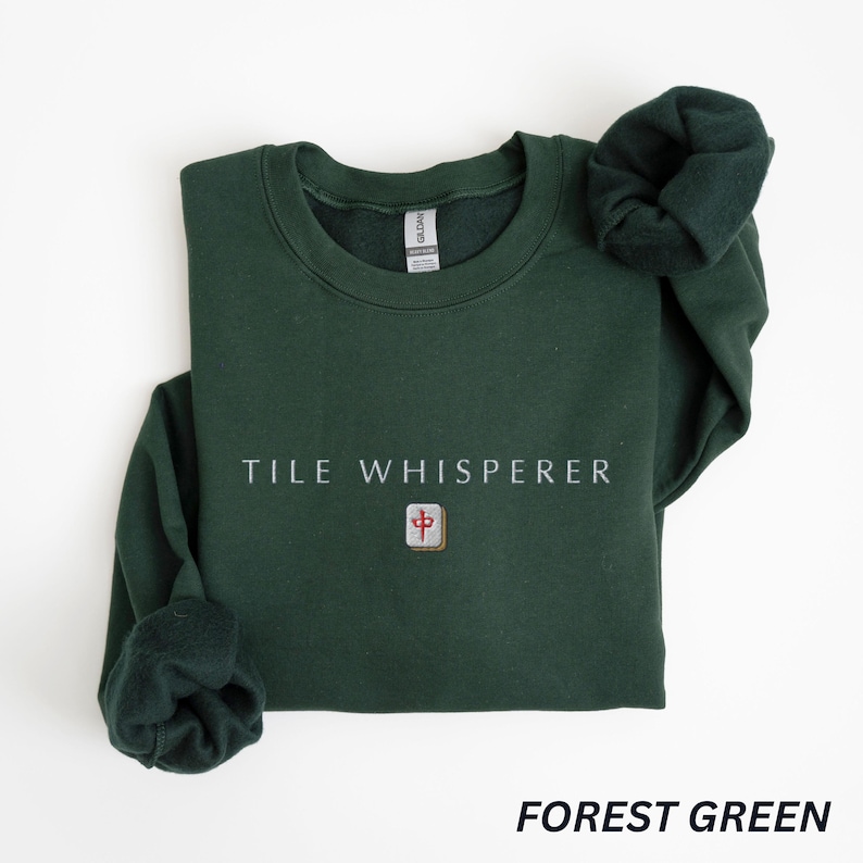 May include: Forest green sweatshirt with the text "TILE WHISPERER" in white. Below the text is a small square graphic with a red and gold design. The sweatshirt is folded with the sleeves visible.