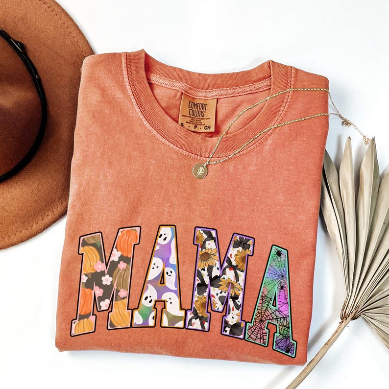 Puede incluir: Una camiseta naranja quemada con la palabra "MAMA" impresa en letras grandes y coloridas. Cada letra est&aacute; llena de un patr&oacute;n diferente con tem&aacute;tica de Halloween, que incluye calabazas, fantasmas, girasoles y telara&ntilde;as.