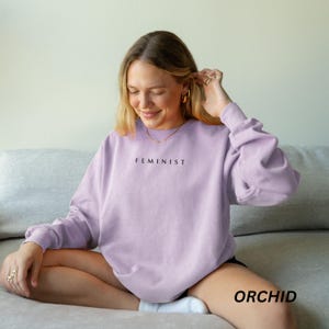 Pu&ograve; includere: Una felpa lilla con la scritta "FEMINIST" stampata in nero. La parola "ORCHID" &egrave; stampata in nero nella parte inferiore della felpa. La felpa &egrave; indossata da una persona seduta su un divano grigio.