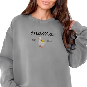 May include: Gray sweatshirt with the word "mama" in cursive script. Below the word "mama" is a small embroidered design of a goose with a yellow chick. The text "EST. 2025" is also included.