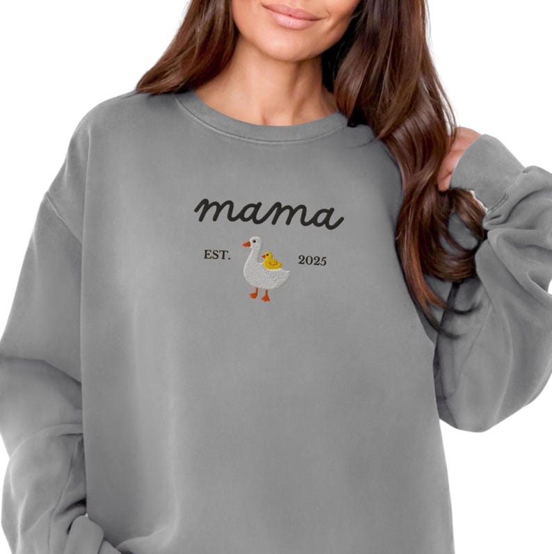 May include: Gray sweatshirt with the word "mama" in cursive script. Below the word "mama" is a small embroidered design of a goose with a yellow chick. The text "EST. 2025" is also included.