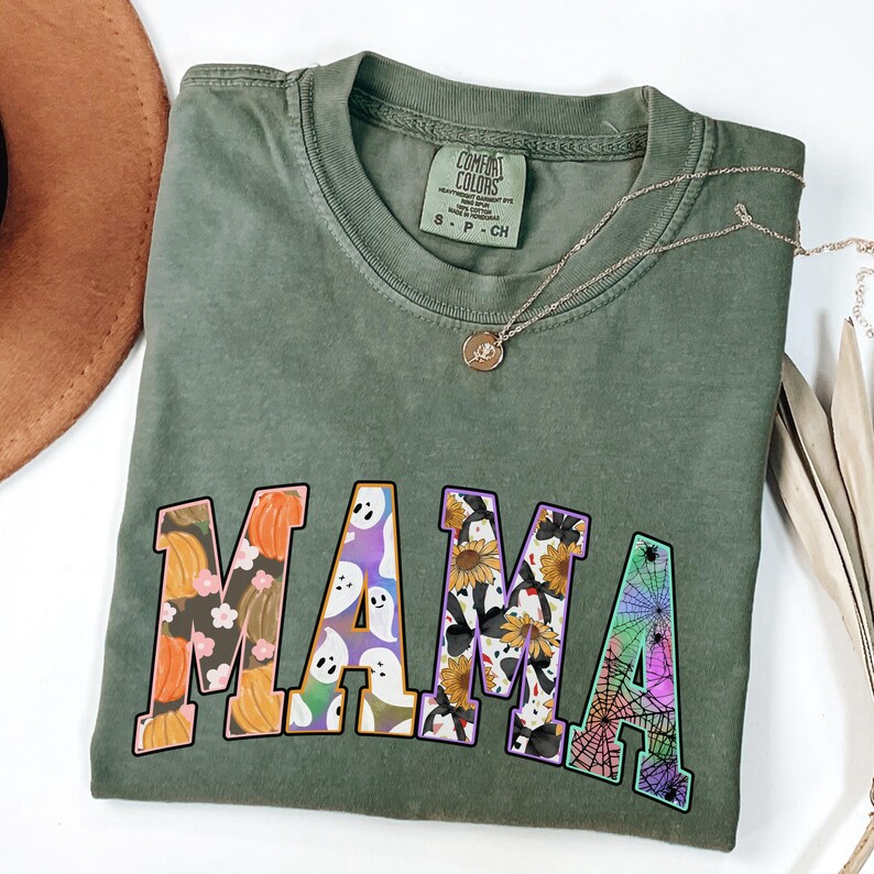Puede incluir: Camiseta verde con un gr&aacute;fico de "MAMA" que presenta una variedad de patrones de tem&aacute;tica de Halloween, incluyendo calabazas, fantasmas, girasoles y telara&ntilde;as.