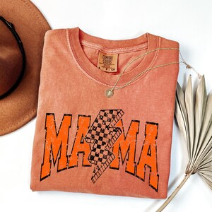 K&ouml;nnte beinhalten: Ein verbranntes orangefarbenes T-Shirt mit einer abgenutzten orangefarbenen Grafik, auf der "MAMA" mit einem karierten Blitz in der Mitte steht. Das Shirt ist gefaltet und auf einer wei&szlig;en Oberfl&auml;che abgelegt.