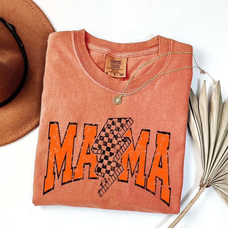K&ouml;nnte beinhalten: Ein verbranntes orangefarbenes T-Shirt mit einer abgenutzten orangefarbenen Grafik, auf der "MAMA" mit einem karierten Blitz in der Mitte steht. Das Shirt ist gefaltet und auf einer wei&szlig;en Oberfl&auml;che abgelegt.
