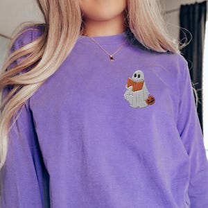 K&ouml;nnte beinhalten: Ein lilafarbenes Langarmshirt mit einem gestickten Geist, der ein Buch und einen K&uuml;rbis h&auml;lt.