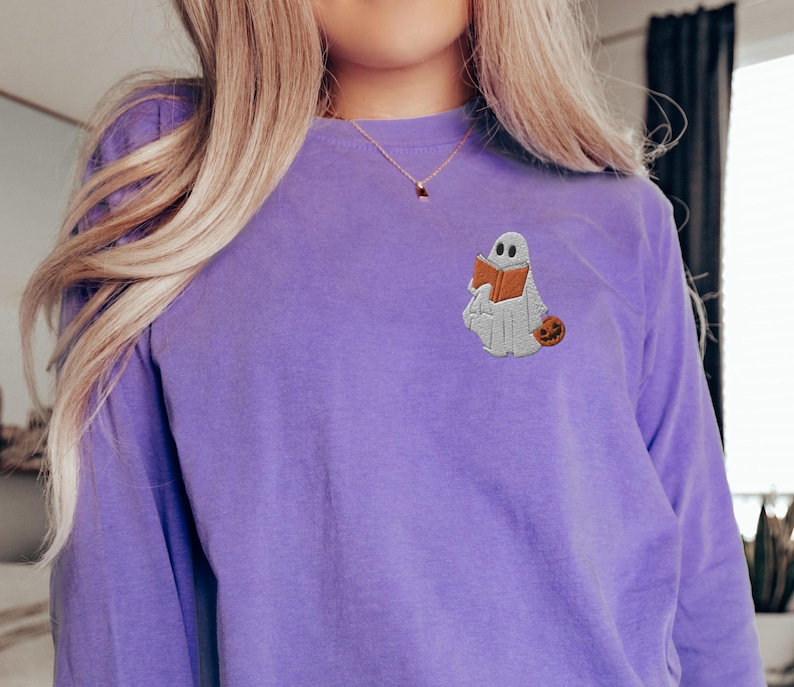 K&ouml;nnte beinhalten: Ein lilafarbenes Langarmshirt mit einem gestickten Geist, der ein Buch und einen K&uuml;rbis h&auml;lt.
