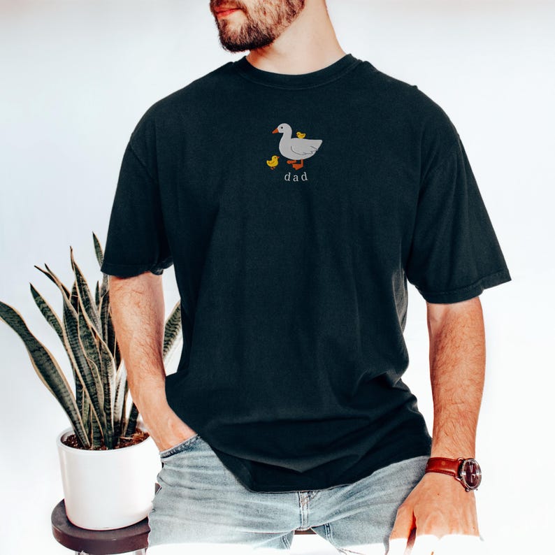 May include: Dark teal t-shirt featuring an embroidered design of a gray duck with two yellow ducklings and the word "dad" in a simple font. The shirt has short sleeves and a relaxed fit.