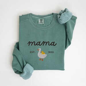 May include: A teal-colored sweatshirt with the word "mama" in a cursive font. Below the word "mama" is an embroidered design of a goose and a duckling, with the text "EST. 2025". The sweatshirt has a crew neck and long sleeves.