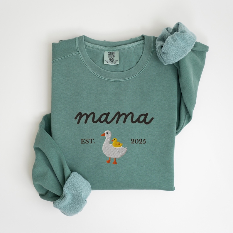 May include: A teal-colored sweatshirt with the word "mama" in a cursive font. Below the word "mama" is an embroidered design of a goose and a duckling, with the text "EST. 2025". The sweatshirt has a crew neck and long sleeves.